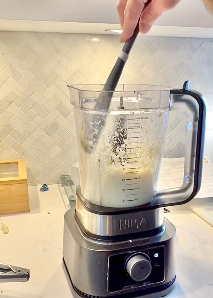 closeup of a hand scraping down the inside of a blender containing the cauliflower