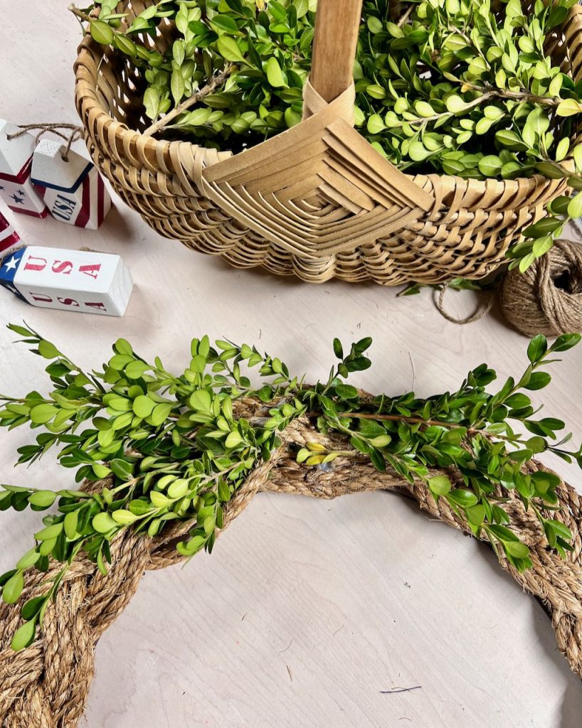tthe top of the braided jute wreath is seen with a basket of fresh boxwood clippings that are being added to the wreath