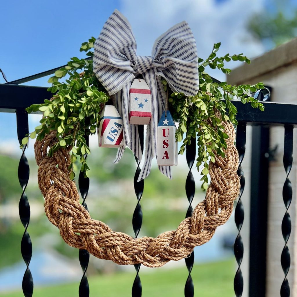 patriotic wreath with navy ticking bow and three wooden buoys is hanging on a black iron gate overlooking a lake