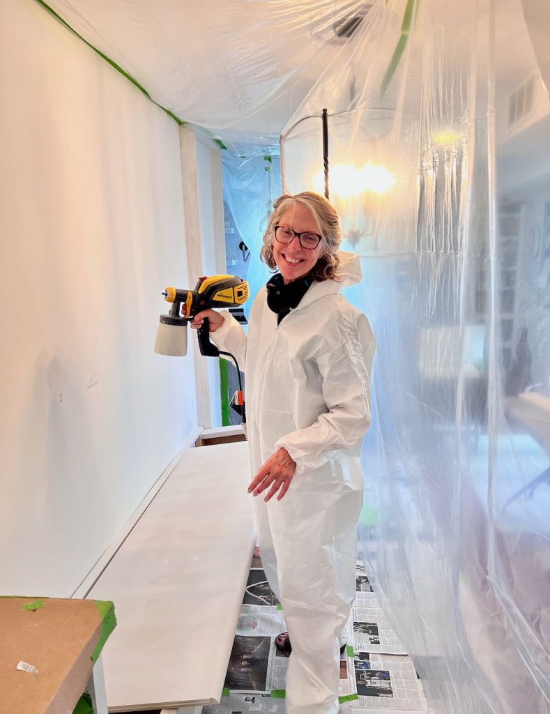 woman in a painting coverall in a room all covered in plastic with a paint sprayer in her hand