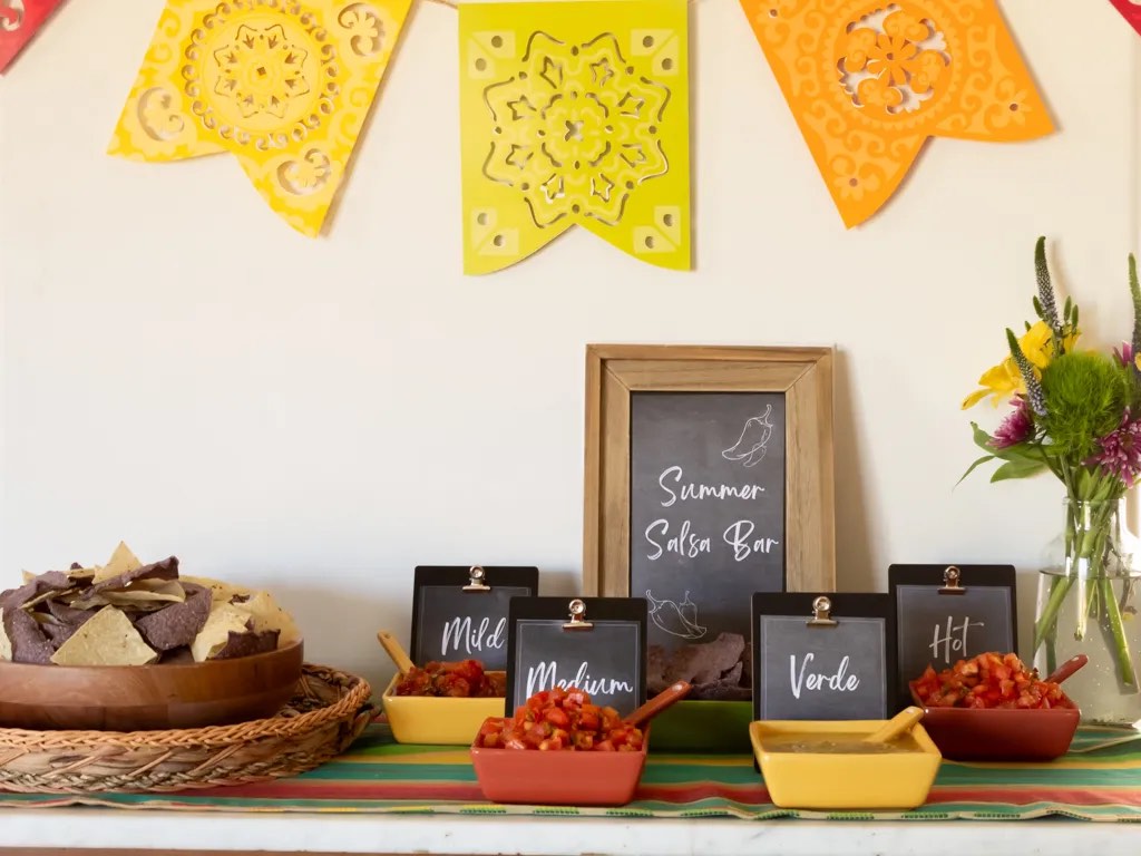Four kinds of salsa in small bowls with chalkboard labels are arranged on a wood cabinet