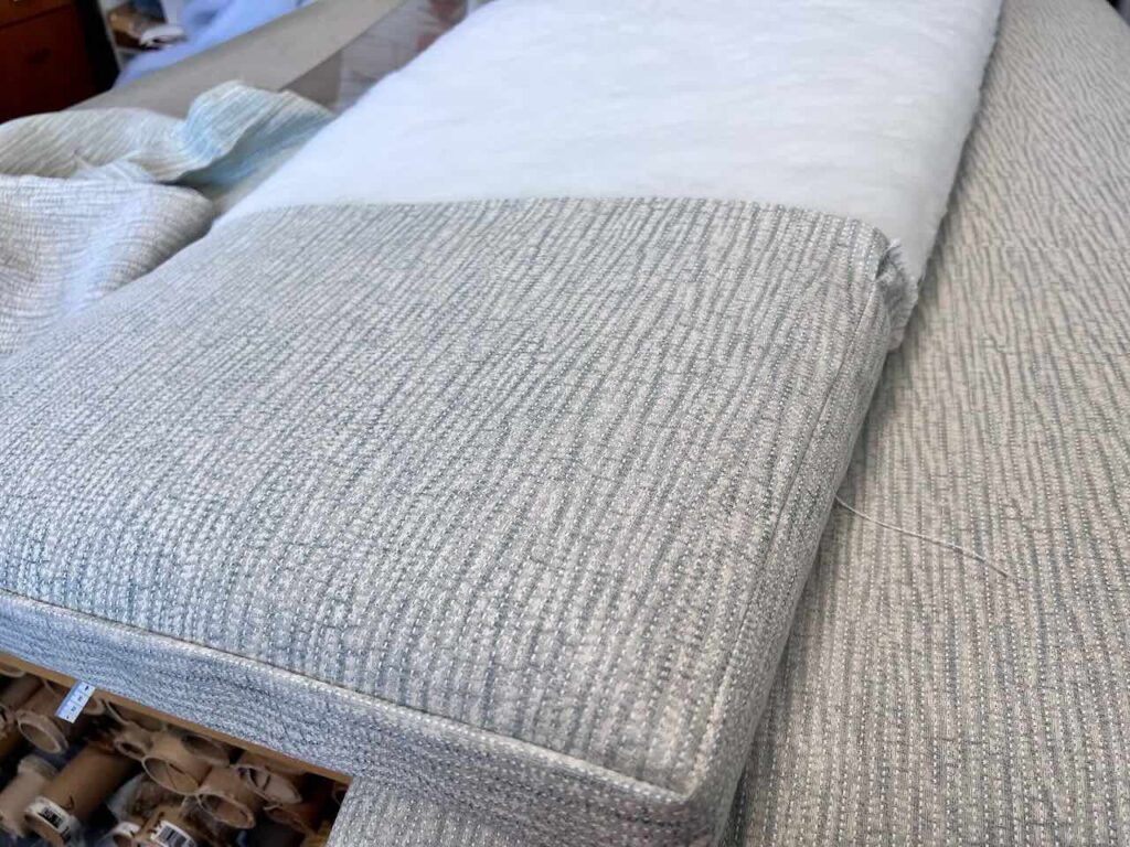 A close-up of an upholstered seat cushion and bench cushion partially covered with batting, showing a textured, light gray fabric. Rolls of similar fabric and an upholstered backrest are visible in the background.
