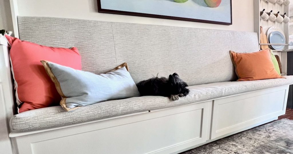 throw pillows and a puppy are lounging on the banquette cushion with an upholstered backrest on the wall