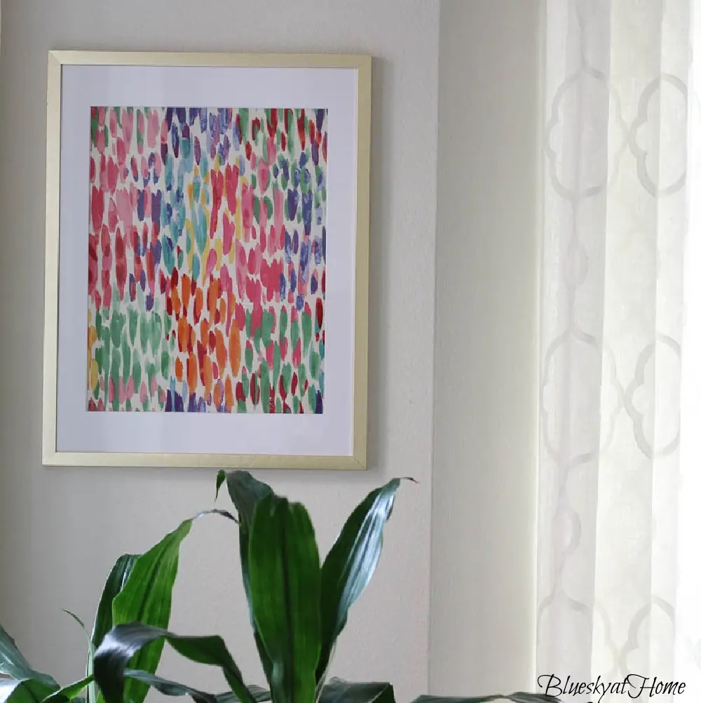 bright colored abstract fabric is in a gold frame hanging beside a window