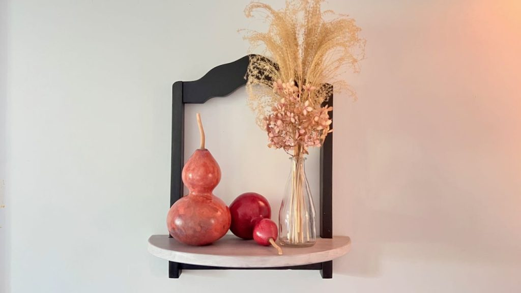 Wide view of Decorative shelf hanging on wall holding a a bottle with dried grasses and hydrangea. next two two dried and dyed gourds