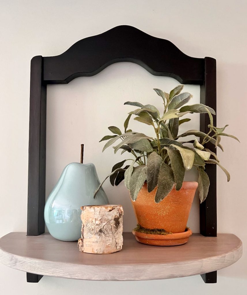 Decorative shelf hanging on wall holding a sage plant in a terra cotta pot next to a ceramic pear and a small. candle in a birch bark holder