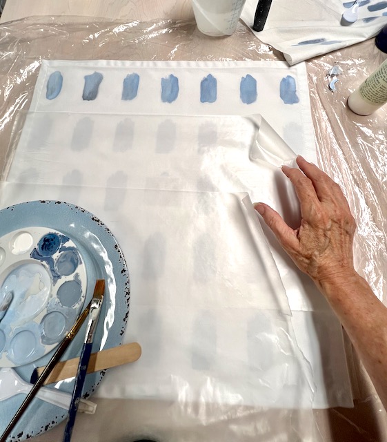 woman's hand arranging two rows of wax paper covering the portion of the napkin that is still to be painted, with a paint palette and brushes on a larger plastic plate
