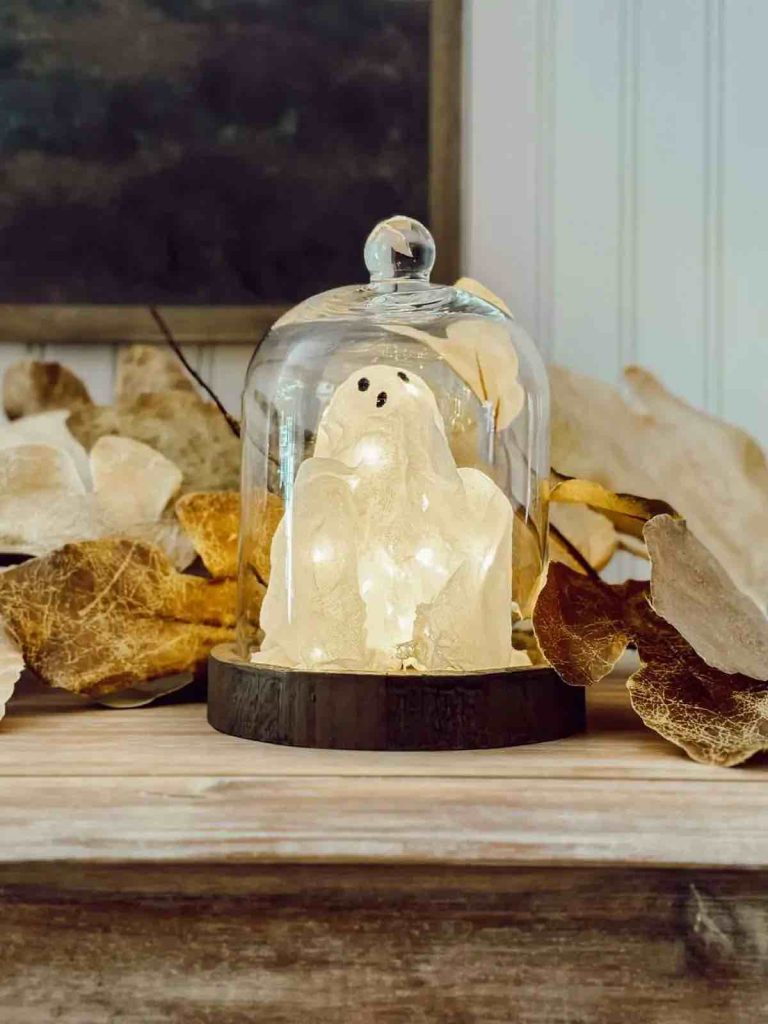 a lit up ghost made of cheesecloth is under a glass cloche surrounded by fall leaves
