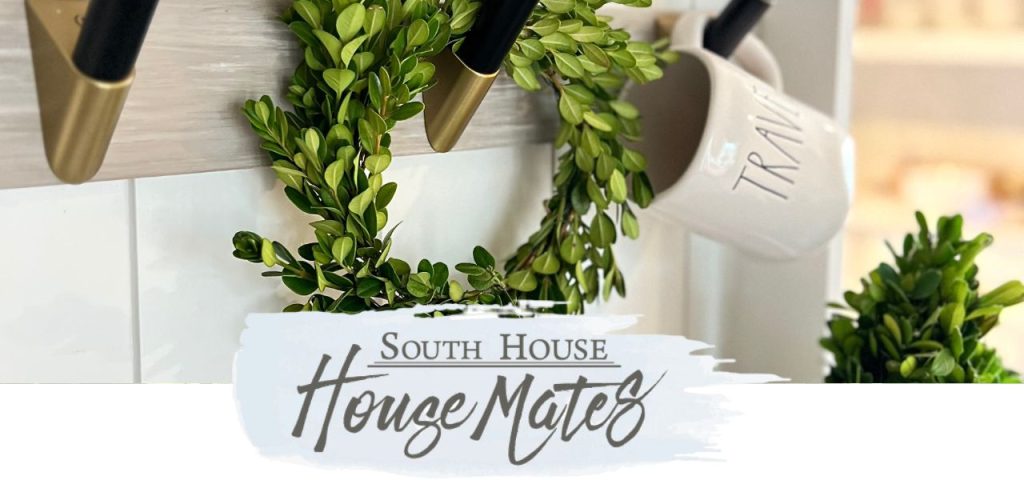 super closeup of ffreshh boxwood wreathh hanging on black and brushed gold hooks next to a mug that says Travel