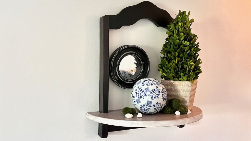 Decorative shelf hanging on wall holding a boxwood tree, ceramic ball and round mirror
