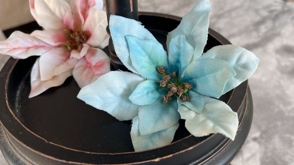 closeup of a finished faux poinsettia flower painted blue resting on a tiered tray with a pink and white flower behind it