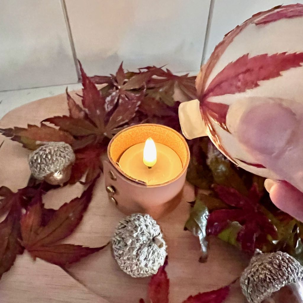 a woman's fingertips are seen holding an ornament decoupaged with Fall leaves being placed over the flameless votive candle