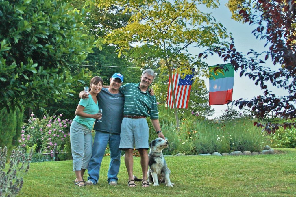 Two men, a woman and dog in a beautiful hillside garden with American and Italian flags hanging behind them
