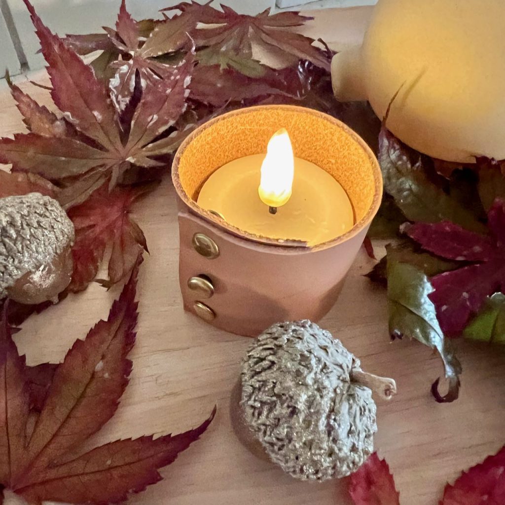 a flameless votive candle is turned on inside a leather sleeve with preserved Fall leaves and acorns around it