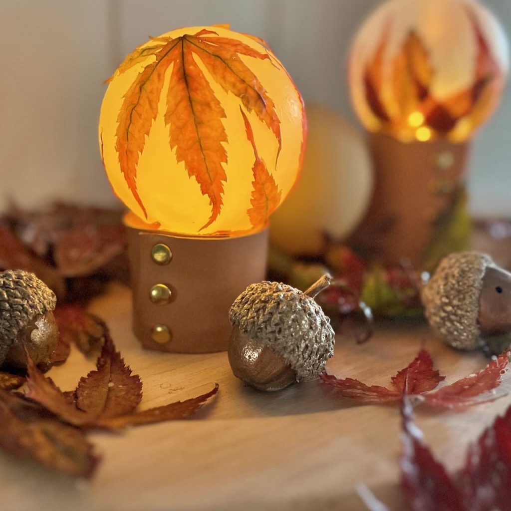 two leaf decoupaged bulbs on leather stands on a wood tray with preserved leaves scattered around
