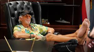 Man in Hawaiian shirt and ball cap in a big leather chair with barefeet  on a desk