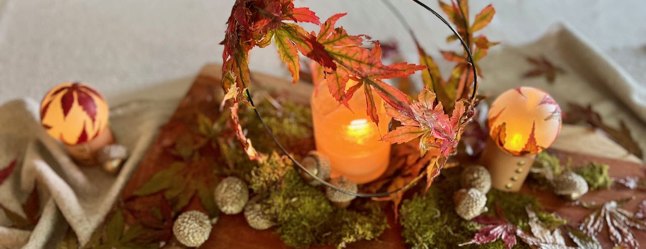 wide closeup of tablescape of Fall leaves and acorns scattered on a linen table runner with three glowing orbs of fall leaves and a large fall leaf orb with candle