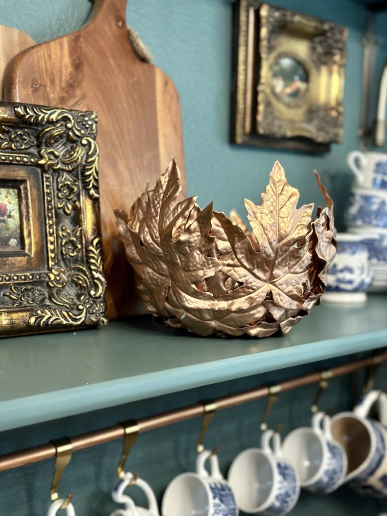 copper leaf bowl in front of a wood cutting board on shelf with cups hanging from brass rod below