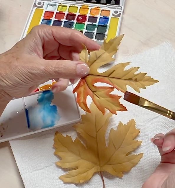 woman's hand is hold a fake leaf while the other hand brush watercolor on it to color the edges
