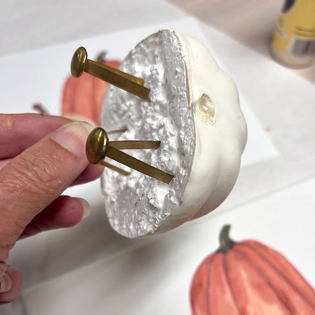 women's fingertips holding onto one of 3 large brads stuck into the styrofoam cut side of a faux pumpkin