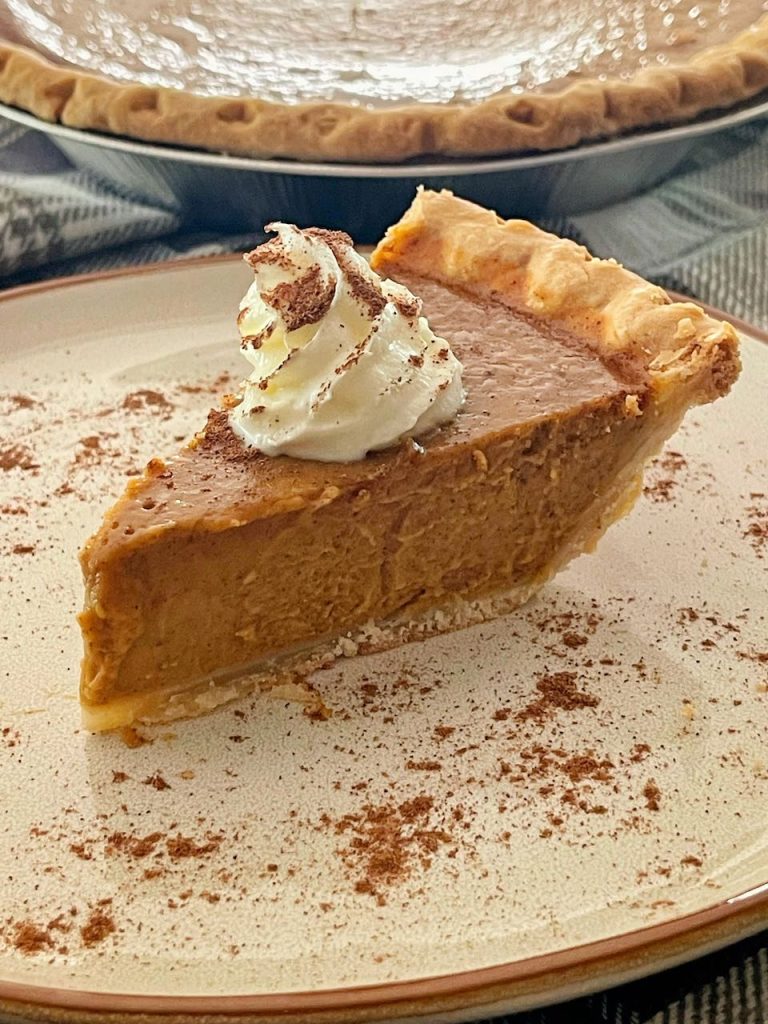 slice of  pumpkin pie is on a plate with a dollop of whipped cream and a dusting of spice