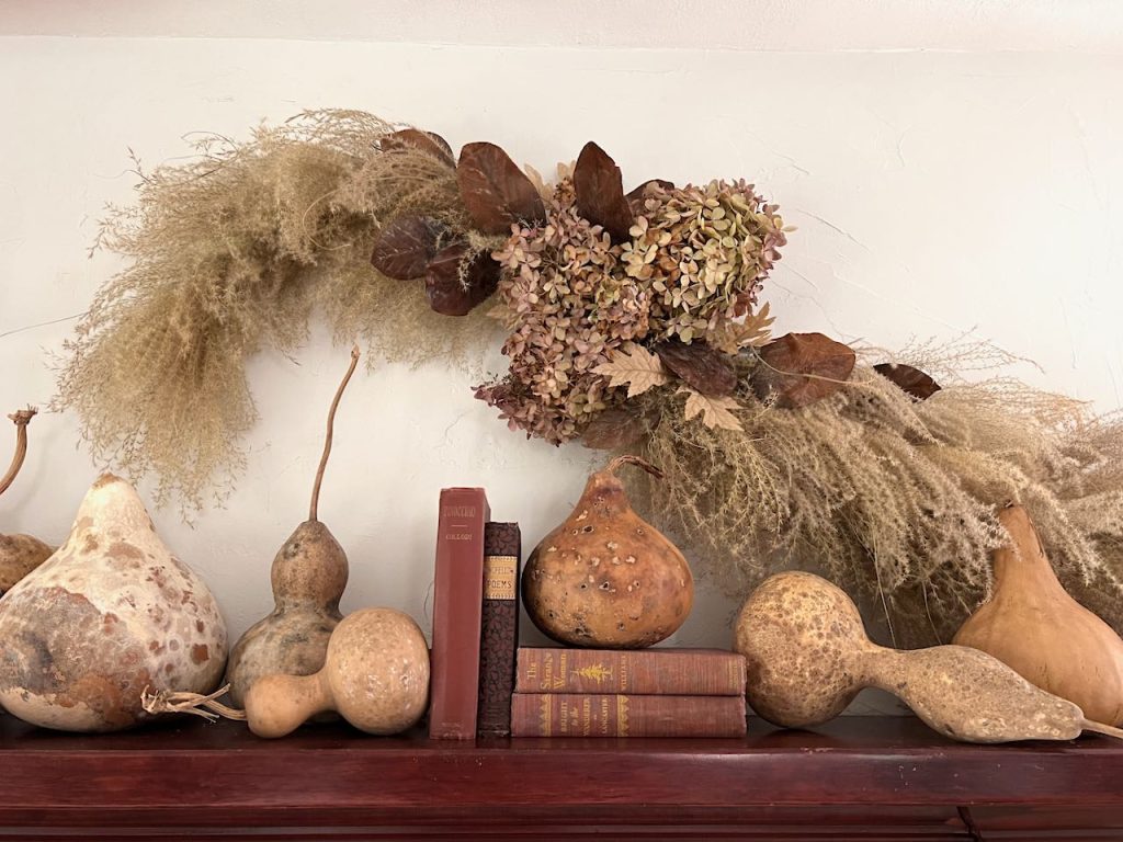 closeup of dried hydrangeas and magnolia leaves with a large garland of dried grass above a mantel full of gourds and old books