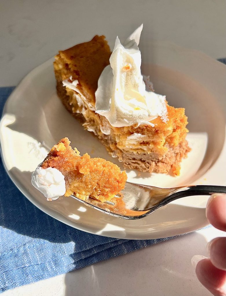 a hand is holding a fork loaded with pumpkin spice gooey butter cake with a bit of whipped cream in front of the rest of the slice on a sunny kitchen counter