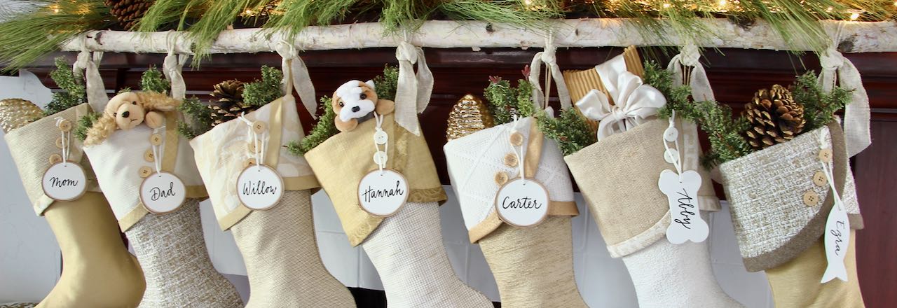 7 coordinating yet unique Christmas stockings are hanging from a birch branch on a mantel above a fire. Tree slice name tags hang from the top of three buttons on each cuff