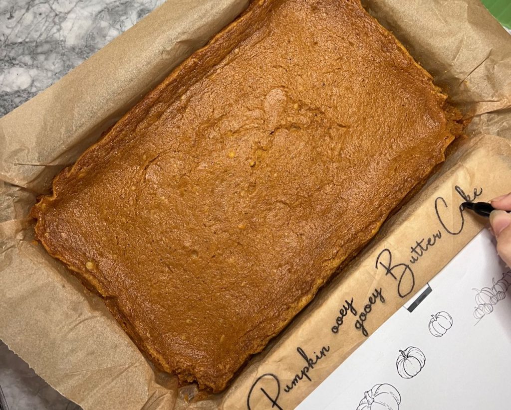 Woman's holding a marker and tracing some words that can be seen through the parchment paper thhat identify the kind of cake in the baking dish