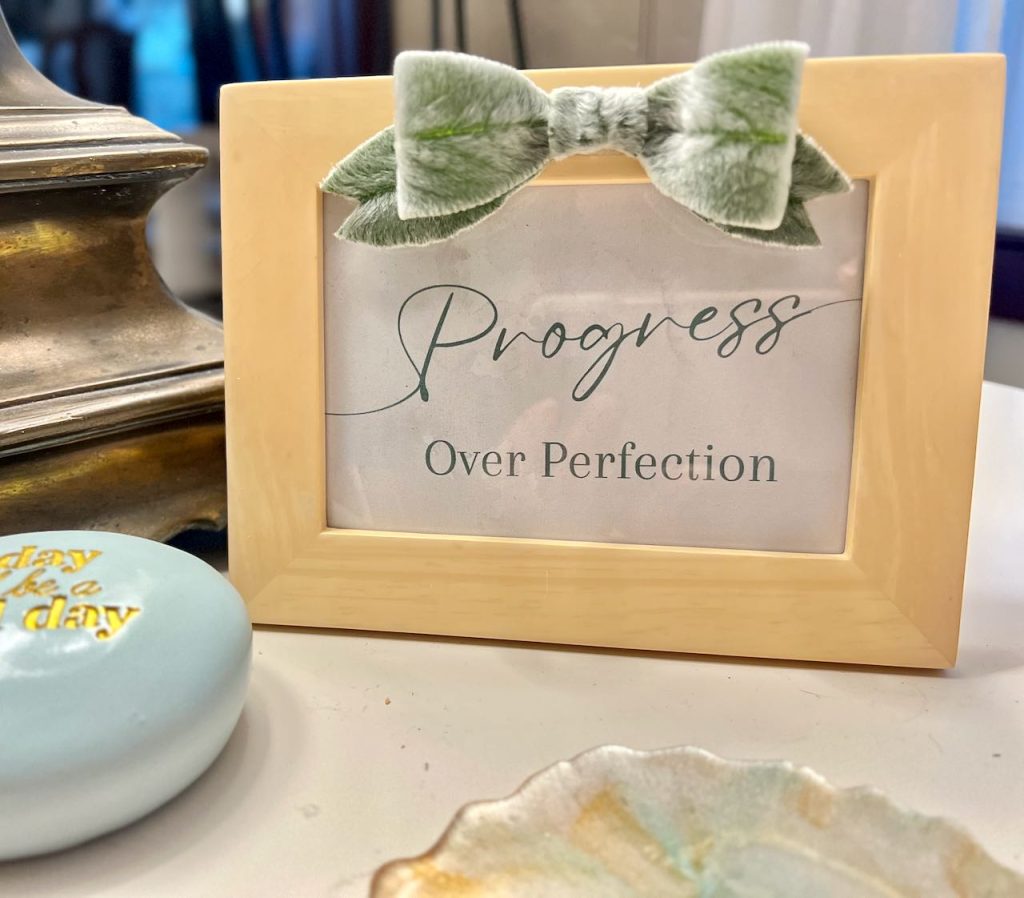 a small frame sitting on a desk holds a reminder "Progress over Perfection" and the frame is embellishhed with a bow made of fresh lambs ear.