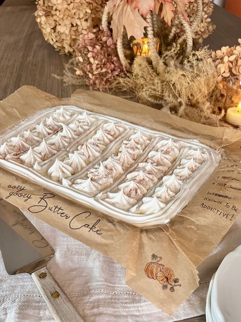 Cake under a cover of whipped cream in a 9 X. 13 baking dish is identified at Pumpkin Ooey Gooey Butter Cake on the Parchment Paper liner that's hanging over the edge