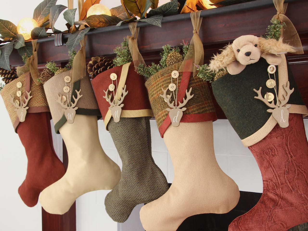 5 coordinating yet unique Christmas stockings are hanging from a mantel. Wood stag head shaped name tags hang from the top of three buttons on each cuff