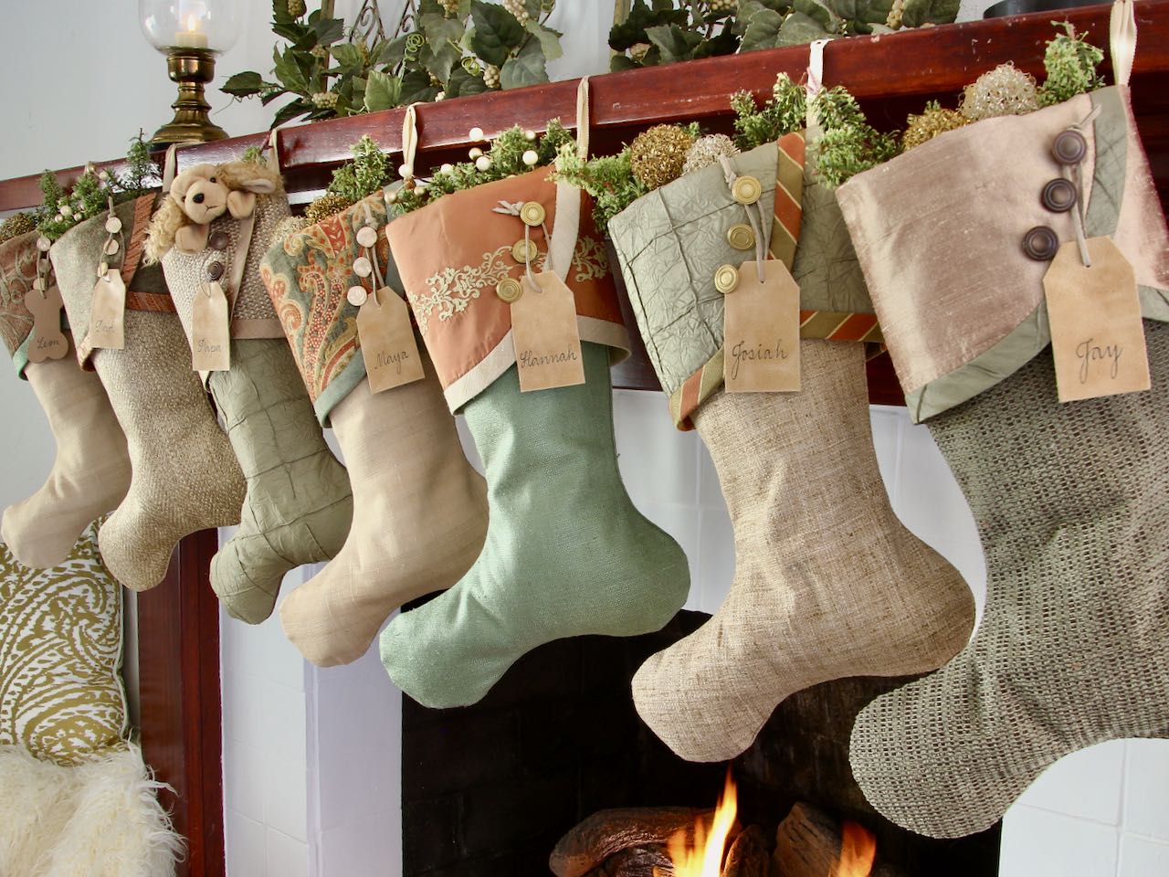 7 coordinating yet unique Luxury Christmas stockings are hagning from a mantel above a fire. Leather name tags hang from the top of three buttons on each cuff
