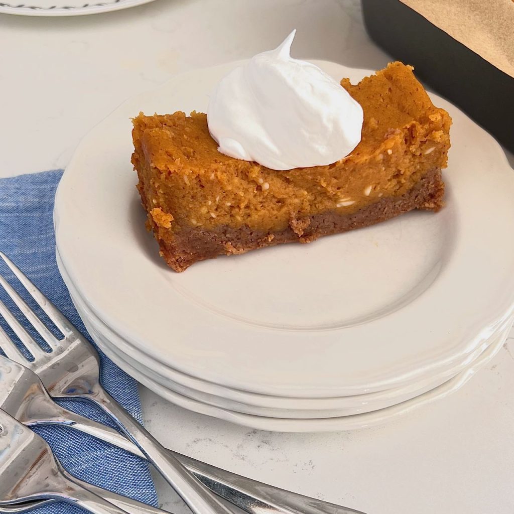Slice of pumpkin spice gooey butter came with a dollop of whipped cream on a stack of plates with forks and napkins nearby