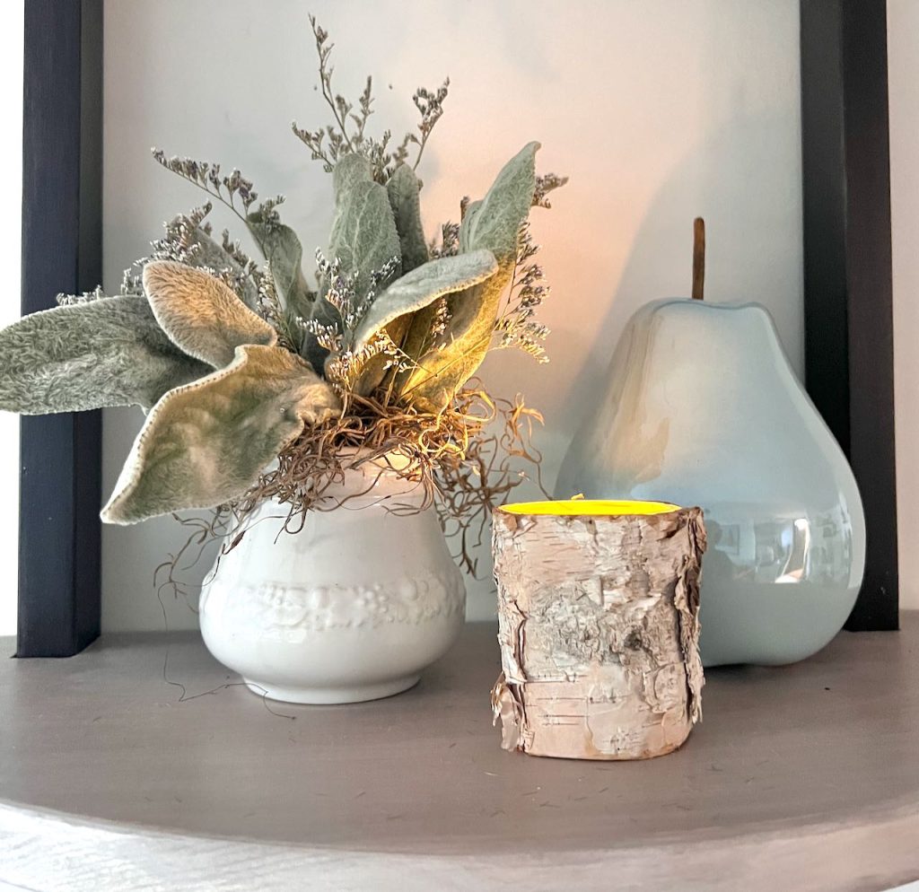 a small arrangement featuring lambs ear is in a sugar dish and sitting on a wall shelf beside votive candle and a ceramic pear