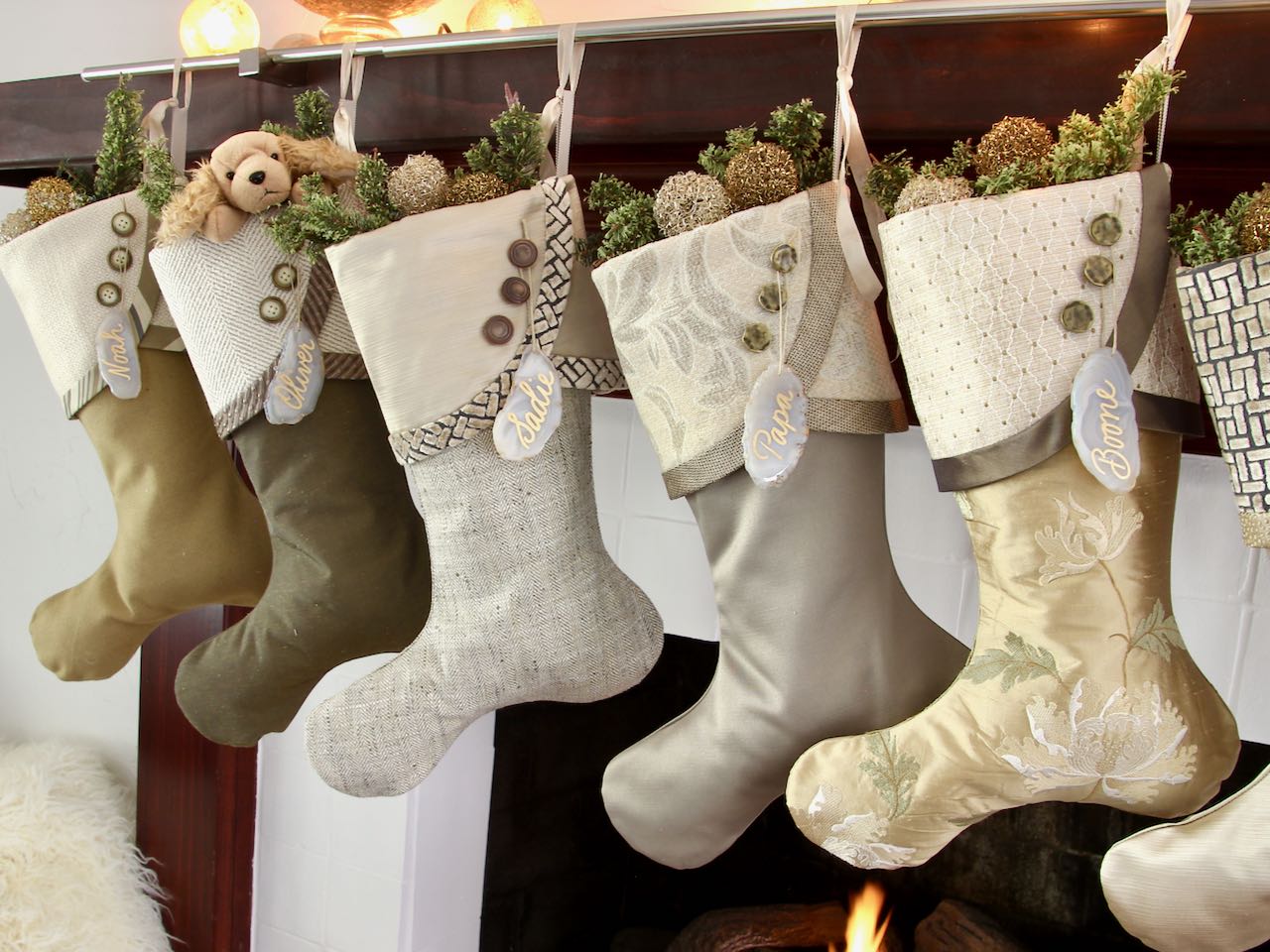 5 coordinating yet unique Christmas stockings are hanging from a mantel above a fire. Agate slice name tags hang from the top of three buttons on each cuff