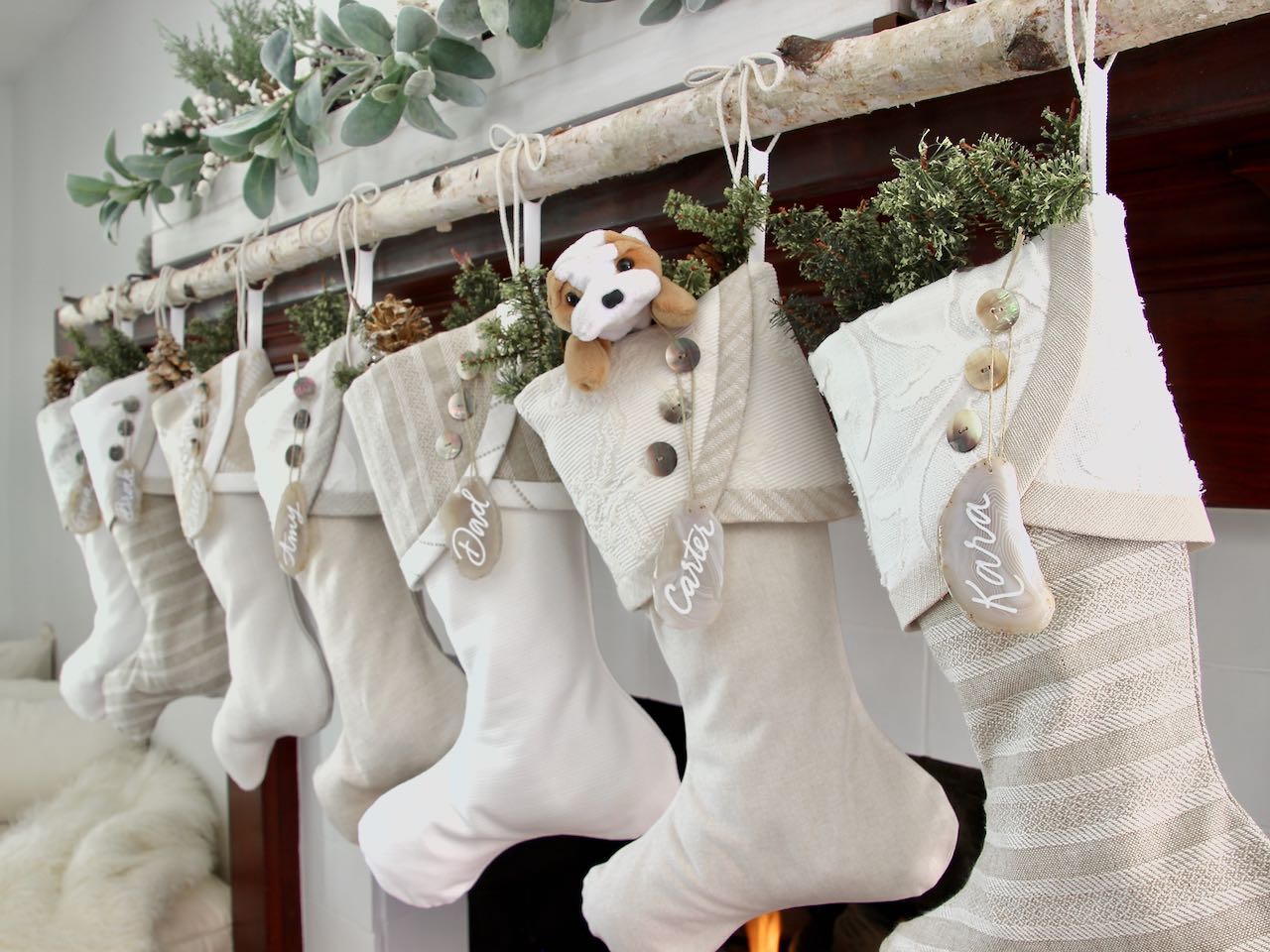 7 coordinating yet unique Christmas stockings are hanging from a mantel above a fire. Agate slice name tags hang from the top of three buttons on each cuff