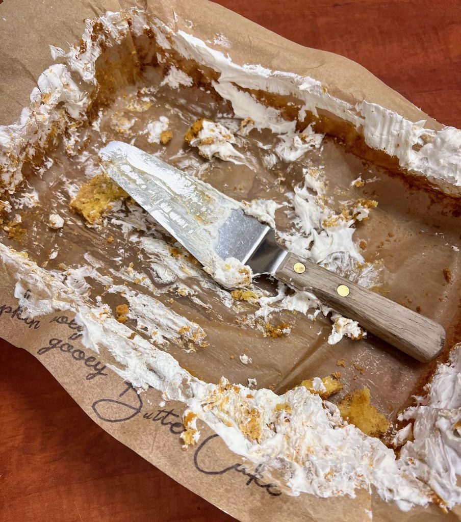 Closeup of a 9 x 13 baking dish with a parchment paper liner that says "Pumpkin Ooey Gooey Butter Cake. All that's left are some crumbs and a bit of whipped cream smeared about