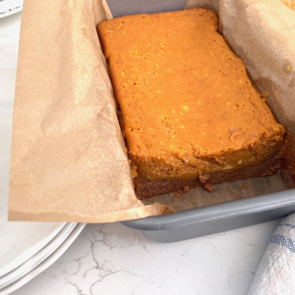 Pumpkin spice ooey gooey butter cake baked in a parchment paper lined loaf pan with one slice missing