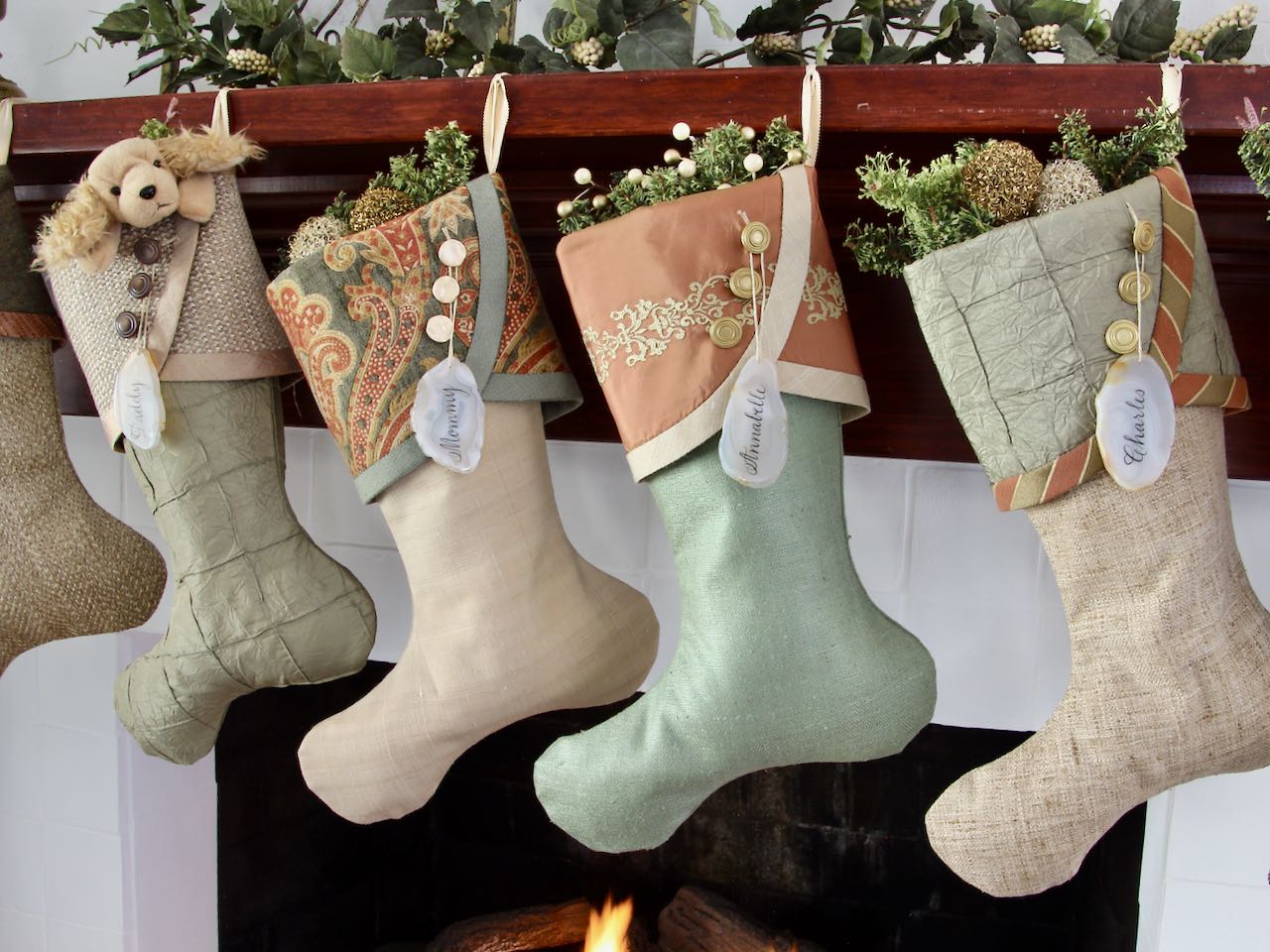 4 coordinating yet unique Luxury Christmas stockings are hanging from a mantel above a fire. agate slice name tags hang from the top of three buttons on each cuff