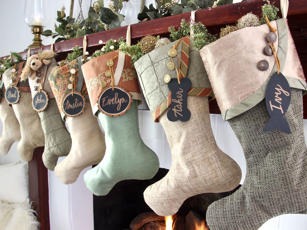 7 coordinating yet unique Luxury Christmas stockings are hagning from a mantel above a fire. Tree slice name tags hang from the top of three buttons on each cuff