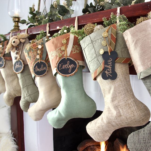 luxe Christmas stockings hang from a mantel with a fire below. Name tags made from tree slices hang from the top button. on each cuff