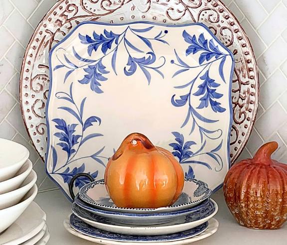 ceramic pumpkin on a pile of small mismatched plates in front of a share plate and a round plate standing in the corner of open shelves