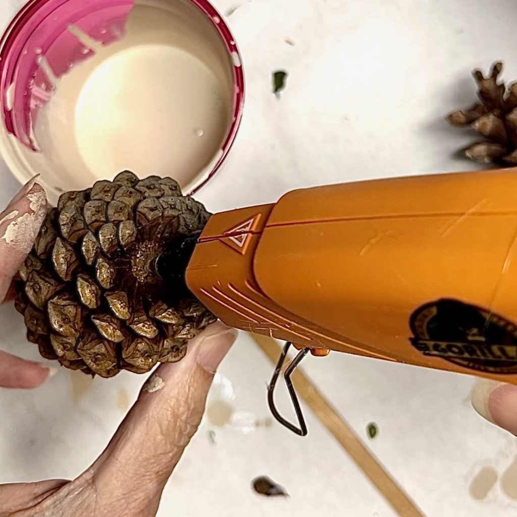 tip of hot glue gun is applying hot glue to the base of a pinecone