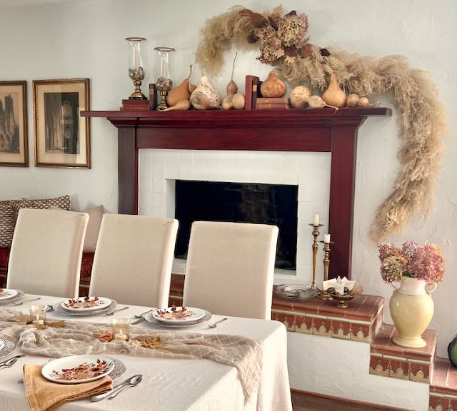 wall-mounted dried arrangement hanging above a mantel with gourds and vintage books. The garland hangs down below thhe mantel on one side with a set dining table in front of the fireplace