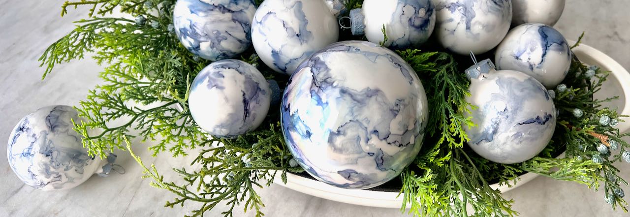 Wide closeup of Christmas bulbs with alcohol ink designs on them are in a bowl of faux Christmas greens