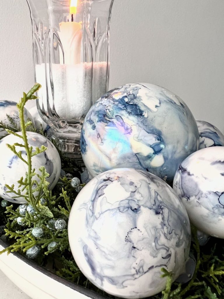 Christmas bulbs with alcohol ink designs on them are in a bowl of faux Christmas greens and a candle in a clear glass hurricane
