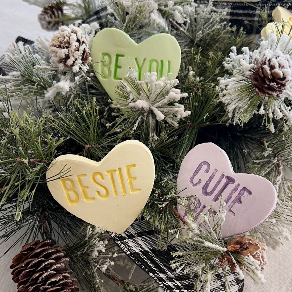 Closeup of large clay conversation hearts that say, BESTIE, BE YOU and  CUTIE PIE nestled in greenery of a centerpiece