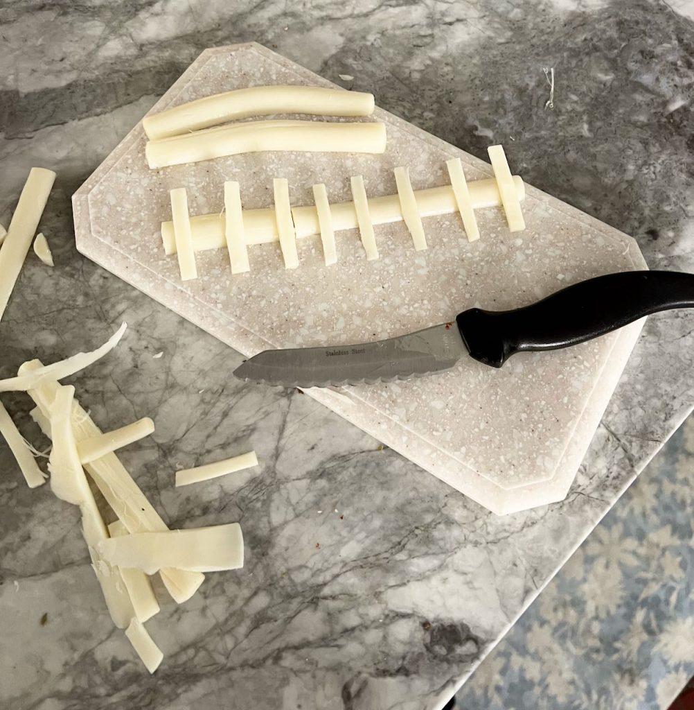 strips of white string cheese are next to a knife and positioned llike the lacing of a football are on a cutting board