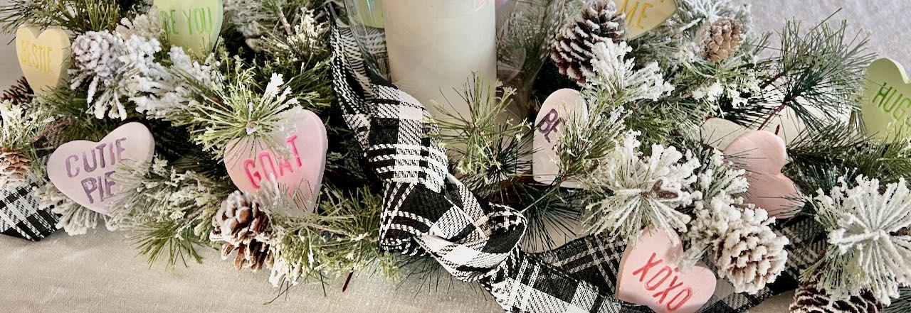 Wide closeup image of a Winter centerpiece with black & white ribbon and large clay conversation hearts nestled in the greenery.